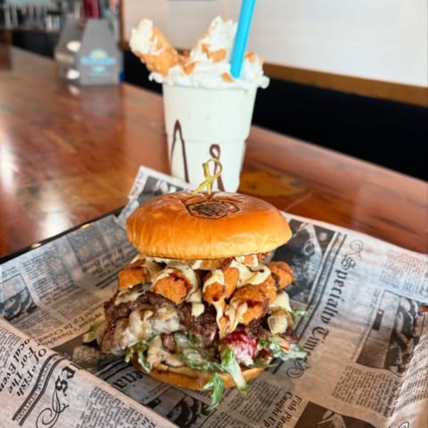A loaded burger with tater tots and sauce, served on newspaper-print paper, with a milkshake in the background.