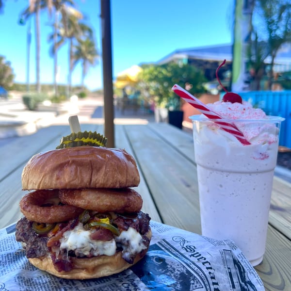 Island Burger and Shake's burger with onion rings and a milkshake on a wooden table outdoors.